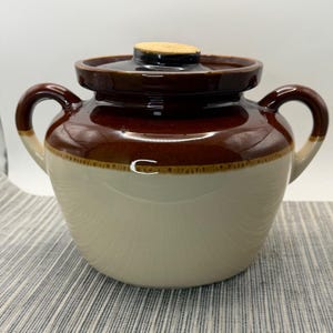 May include: A two-handled ceramic crock featuring a glossy dark brown glaze on the upper portion and a smooth cream-colored glaze on the lower body. The vessel includes a matching dark brown lid with a small, light-colored knob at the center.
