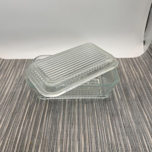 May include: A rectangular clear glass butter dish with a matching lid. The lid features a series of parallel vertical ridges across its surface, while the base has a corresponding ribbed texture along its sides, creating a uniform design.