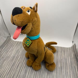 May include: A brown plush dog toy with a large black nose, a pink tongue sticking out, and a bright turquoise collar featuring a yellow diamond-shaped tag with the black letters SD printed on it. The plush has dark brown spots on its back.