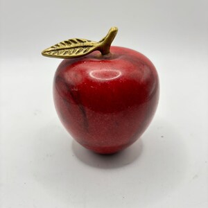 May include: A decorative red apple figurine with a gold-colored leaf and stem. The apple has a glossy finish and a marble-like texture. The leaf is detailed with etched veins.