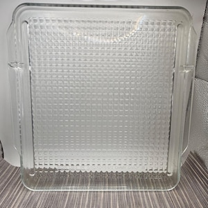 May include: A clear, square glass baking dish featuring a textured grid pattern on the base. The dish includes integrated side handles and the text FOR USE IN MICROWAVE OVEN ONLY embossed along the bottom edge of the interior surface.