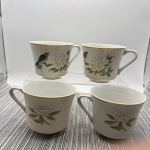 May include: A set of four white porcelain teacups featuring gold-trimmed rims. Each cup is decorated with botanical illustrations of white flowers and green leaves, with two cups also displaying a small bird perched on a branch.