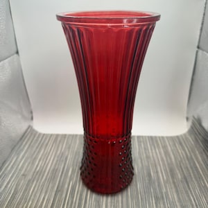 Peut inclure: Un grand vase en verre rouge translucide doté d'un bord évasé et large ainsi que d'un corps cylindrique. La partie supérieure du vase est ornée de lignes verticales nervurées, tandis que la section inférieure présente un motif à pois en relief.