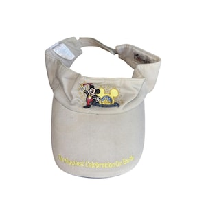 May include: Tan visor featuring an embroidered image of Mickey Mouse in a celebratory outfit. The visor has the text "The Happiest Celebration On Earth" in yellow. Adjustable strap in the back.
