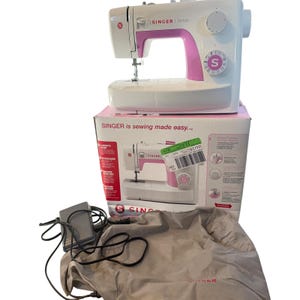 May include: A white and pink Singer Simple sewing machine sits atop its original cardboard box. The box features the text SINGER is sewing made easy. A grey foot pedal with a black power cord and a grey fabric dust cover are included.