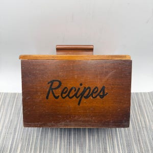 May include: A rectangular wooden recipe box with a dark brown finish and a hinged lid. The word Recipes is printed in black cursive script on the front panel. The box features a smooth, flat top handle and shows natural wood grain textures.