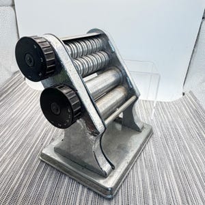 May include: A silver-toned metal pasta machine featuring two sets of rollers and two black circular adjustment knobs on the side. The top knob is numbered one through four, while the bottom knob is numbered one through six for thickness control.