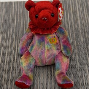 May include: A plush teddy bear with a solid red head and a red ruffled collar. The body features a multi-colored tie-dye pattern in shades of blue, pink, yellow, and green. An embroidered patch on the chest reads JULY with candles.