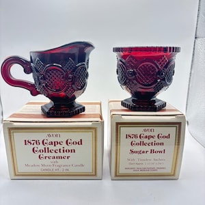 May include: A ruby red glass creamer and sugar bowl set from the Avon 1876 Cape Cod Collection. The creamer has a handle and spout, while the sugar bowl is a pedestal design. Both are on their original boxes, which include the text "Meadow Morn Fragrance Candle" and "Sugar Bowl".