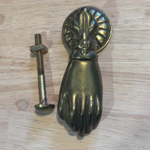 May include: A vintage brass door knocker in the shape of a hand, with a decorative circular backplate. A matching brass bolt is included. The knocker has a detailed design and a warm, aged patina, suggesting it is an antique.