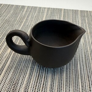 May include: A matte black ceramic creamer or small pitcher with a rounded body and a curved handle. The vessel features a distinct pouring spout on the rim, designed for liquid service. The surface has a uniform dark finish with a smooth texture.