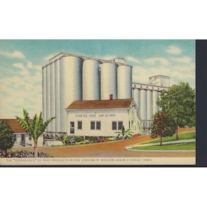 May include: Vintage postcard featuring the "birthplace" of Post Products. The image shows a white building with the text "STARTED HERE JAN 1ST 1895" in front of large grain storage tanks. The scene is set under a blue sky with some clouds.
