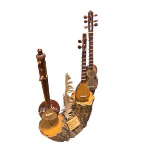 May include: A wooden sculpture featuring three miniature stringed instruments, a feather, and a small plaque. The instruments are brown with intricate details, mounted on a section of a tree trunk. The plaque has text in a foreign language.
