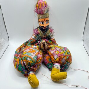 May include: A colorful, handcrafted puppet with a painted wooden face, black mustache, and beard. The puppet wears a patterned fabric outfit in shades of pink, orange, green, and yellow. The puppet's legs are yellow, and it has a decorative headpiece.