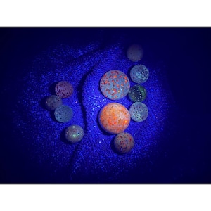 May include: A collection of glowing, spherical objects in various colors, including orange, red, and blue, are arranged on a dark blue, textured surface. The spheres have intricate patterns and textures, creating a visually striking display. The image is lit with a blue light.
