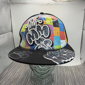 Peut inclure: Une casquette de baseball noire avec un motif à grille multicolore et le texte Mickey Mouse répété en plusieurs couleurs. Un grand contour de tête de Mickey Mouse brodé en blanc est centré sur le devant, avec le texte Mickey Mouse sur la visière.