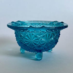 May include: A small, turquoise-colored glass bowl with a diamond pattern and three small feet. The rim of the bowl has a scalloped edge. The bowl is likely vintage and is suitable for serving small portions.