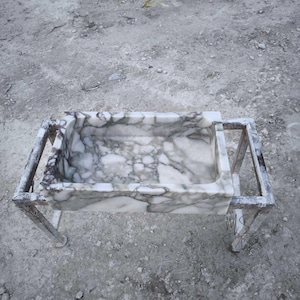 May include: A white marble sink with gray veining, resting on a metal frame. The sink is rectangular and has a slightly rounded edge. The frame is made of metal bars and is sturdy.