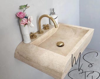 May include: Beige stone sink with a curved design, featuring gold-colored faucet and fixtures. A white marble vase with pink and white flowers sits on the side. A soap dispenser is also present. The sink has a modern, minimalist aesthetic.