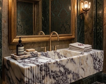 May include: A marble bathroom sink with a fluted design, featuring white and purple veining. The sink has gold taps and is set against a backdrop of green and gold decor. The mirror is framed in gold.