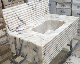 May include: A white marble bathroom sink with a rectangular basin and a back splash. The sink has a ribbed design on the front and sides, with dark veining throughout the marble. The sink is on a metal stand.