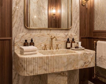 May include: A luxurious bathroom sink made of beige marble with a fluted design. The sink has a gold faucet and is surrounded by a marble backsplash and wooden paneling. Soap dispensers and towels are visible.