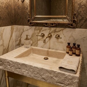 May include: A beige stone sink with a textured edge, supported by a gold-coloured metal frame. A gold-framed mirror hangs above the sink, reflecting the patterned wallpaper. A glass soap dispenser and a folded towel are on the sink.
