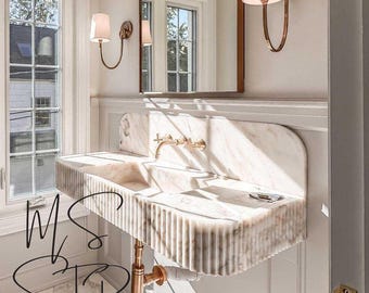 May include: A white marble bathroom sink with a rippled edge and a gold faucet. The sink is mounted on a wall and has a gold drain pipe.