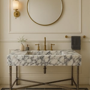 Peut inclure: Une vasque de salle de bain en marbre blanc avec un robinet en laiton et un miroir rond au-dessus. La vasque a une base en laiton avec un motif en forme de croix.