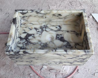 May include: Rectangular marble sink with a white and black veined pattern. The sink has a central drain hole and is made of a polished stone material. The sink is a bathroom fixture.