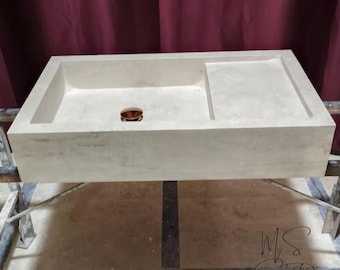 May include: A rectangular, light grey concrete bathroom sink with a built-in shelf on the right. The sink has a single drain hole with a copper-coloured ring. The sink is supported by a metal frame, with a maroon curtain in the background.