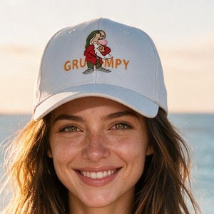 May include: A white baseball cap featuring an embroidered graphic of the character Grumpy from Snow White and the Seven Dwarfs, with the word GRUMPY printed in orange letters underneath the character illustration on the front panel.