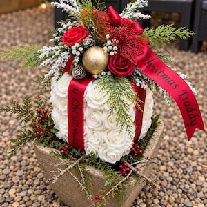 Może przedstawiać: Kwadratowa donica wypełniona kostką z białych sztucznych róż, owiniętą czerwoną wstążką z napisem Merry Christmas. Kompozycję wieńczą czerwone róże, złota bombka, szyszki, gałązki cedru oraz czerwona wstążka z napisem Christmas Daddy.