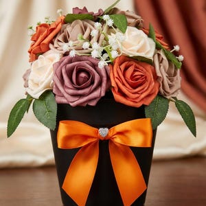 May include: A floral arrangement in a black pot with an orange bow. The bouquet features roses in shades of orange, cream, and burgundy, with green leaves and small white flowers. A small, heart-shaped jewel is in the center of the bow.