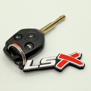 Puede incluir: Una llave de coche negra con un llavero y un llavero de metal. El llavero tiene botones para varias funciones. Un llavero LSX negro, blanco y rojo está unido al llavero.