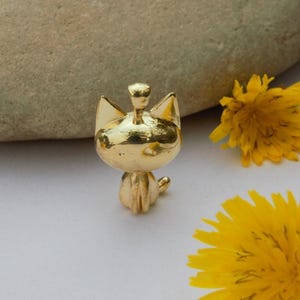 Puede incluir: Un amuleto de metal dorado en forma de gato sentado sobre sus patas traseras. El amuleto está sobre una superficie blanca con flores amarillas.