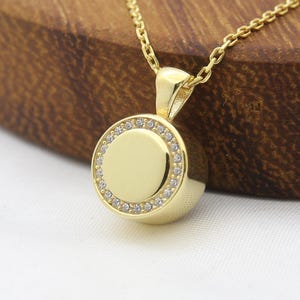 May include: A gold pendant necklace featuring a circular charm. The charm is embellished with small, clear stones. The necklace and pendant are displayed on a wooden surface, against a white backdrop.