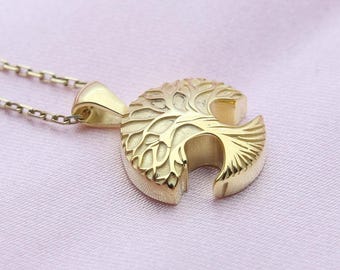 May include: A gold-colored pendant necklace featuring a tree of life design. The pendant is a half-circle shape with intricate detailing. The necklace chain is delicate and also gold-colored. The background is a soft pink fabric.