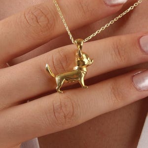 Op de afbeelding: Een goudkleurige hangerketting met een kleine, gedetailleerde chihuahua-hondenbedel. De bedel hangt aan een delicate ketting. De ketting wordt getoond op een wit overhemd.