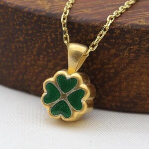 May include: A gold-colored necklace with a four-leaf clover pendant. The clover is green with a gold border. The necklace chain is delicate and gold, and the pendant is centered. The necklace is displayed on a wooden surface.