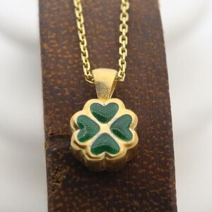 May include: A gold necklace with a four-leaf clover pendant. The clover is green with a gold outline. The necklace is displayed on a brown surface.