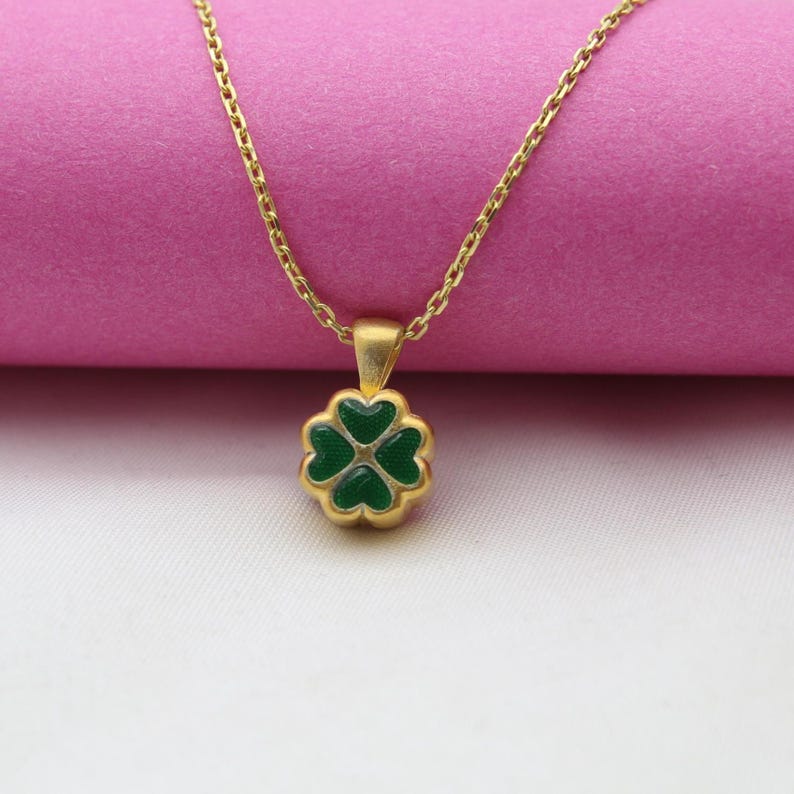 May include: A gold-colored necklace with a four-leaf clover pendant. The clover is green with a gold border, and the chain is delicate. The necklace is displayed against a pink and white background.