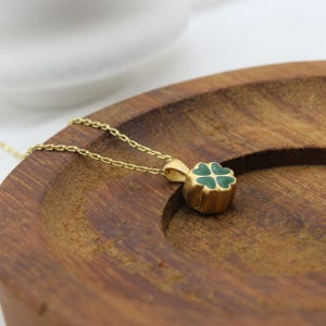 May include: A gold-colored necklace with a four-leaf clover pendant. The clover is green with gold-colored outlines. The necklace is resting on a wooden surface.