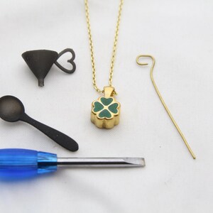 May include: A gold-colored necklace with a four-leaf clover pendant with green enamel. Also, a small black funnel, a black spoon, a blue-handled screwdriver, and a gold-colored wire are arranged on a white surface.