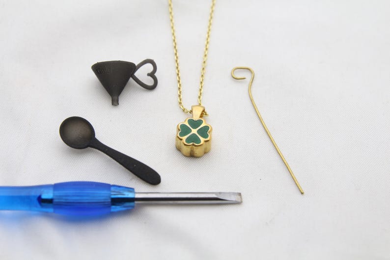 May include: A gold-colored necklace with a four-leaf clover pendant with green enamel. Also, a small black funnel, a black spoon, a blue-handled screwdriver, and a gold-colored wire are arranged on a white surface.