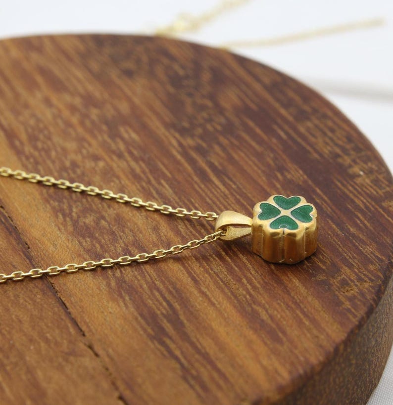 May include: A gold-colored necklace with a four-leaf clover pendant. The clover is green and set in a gold-colored frame. The necklace chain is delicate and gold-colored. The pendant is approximately 0.5 inches in diameter.