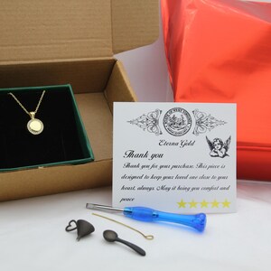 May include: A gold-colored necklace with a circular pendant, presented in a box with a green interior. The box is open, revealing the necklace. A card with text and a red package are also visible.