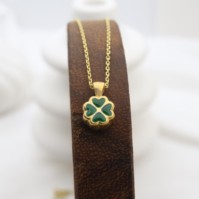 May include: A gold necklace with a four-leaf clover pendant. The clover is green with a gold outline. The necklace is displayed on a brown surface.