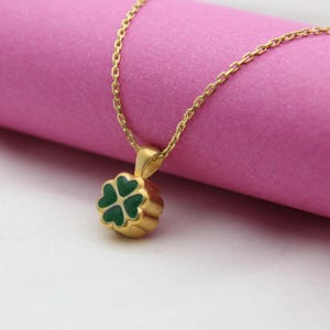May include: A gold-colored necklace with a four-leaf clover pendant. The clover is green with gold trim, and the chain is delicate. The necklace is displayed on a pink surface.