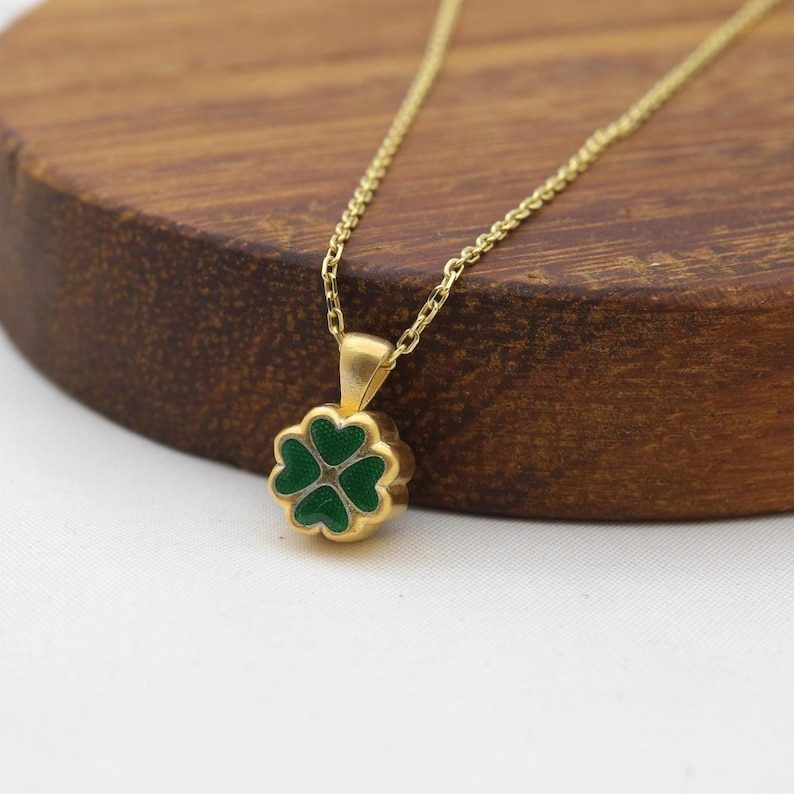 May include: A gold-colored necklace with a four-leaf clover pendant. The clover is green with a gold border. The necklace chain is delicate and gold, and the pendant is centered. The necklace is displayed on a wooden surface.